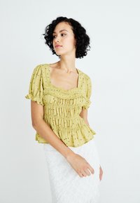 Groen-geel kortgeroffelde blouse met een vierkante halslijn, gesmokt lijfje, hierin ruffled mouwen en verfijnde bloemmotieven.