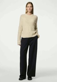 Bežs adīts džemperis ar apaļu kakla izgriezumu, garām piedurknēm un ribota struktūra, apvienots ar platu kāju tumši zilām biksēm un melniem apaviem.