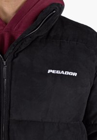Schwarze Jacke mit Reißverschluss und weißem „PEGADOR“-Logo auf der Brust, getragen über einem roten Kapuzenpullover.