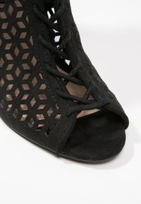 Zapato negro de punta abierta con un patrón hexagonal recortado, material de ante y cordones negros. El tacón también tiene textura con un interior suave.