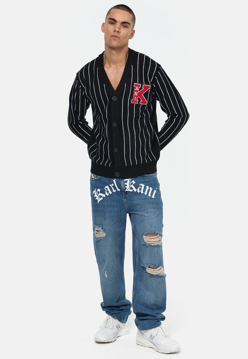 Svart stripet cardigan med et rødt bokstav K, med knappelukking. Brukt sammen med lyseblå slitne jeans og hvite joggesko.
