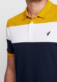 Homme portant un polo à manches courtes avec des blocs de couleur horizontaux moutarde, blanc et bleu marine, et un petit logo d'oiseau sur la poitrine.