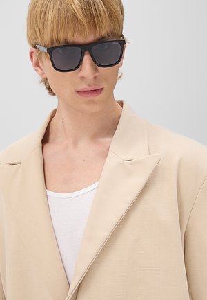 Giovane con capelli biondi corti che indossa occhiali da sole neri, un blazer beige e una maglietta bianca su uno sfondo uniforme.