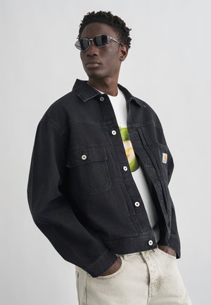 Mężczyzna w czarnej kurtce dżinsowej, białej koszulce z nadrukiem, jasnobeżowych spodniach i małych czarnych okularach przeciwsłonecznych, pozujący z rękami w kieszeniach.