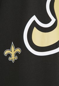 Černá látka s velkým bílým a zlatým grafickým designem, obsahující stylizované písmeno a zlatý emblém lilie v levém dolním rohu.
