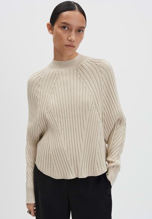 Woman with dark hair tied back wearing a beige ribbed knit sweater with a high neckline and black pants against a neutral background.
