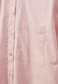Svijetloružičasta košulja na dugmad izrađena od glatkog materijala, s jednom džepom na prsima i suptilnim detaljem šavova duž ruba džepa.