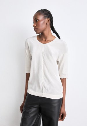 Light beige short-sleeve top with a round neckline, textured fabric, and a central seam detail, paired with black leather trousers.