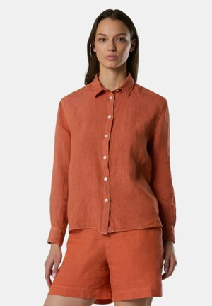 Donna che indossa una camicia color ruggine a maniche lunghe con bottoni e pantaloncini abbinati, in piedi contro uno sfondo chiaro e semplice.