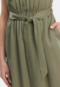 Abito verde oliva con chiusura a bottoni sul davanti, gonna plissettata e cintura legata in vita. Tessuto morbido e senza motivi visibili.