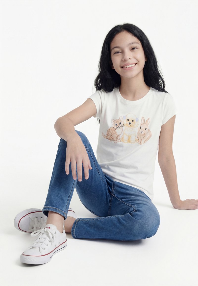 Κορίτσι που χαμογελά με μακριά μαύρα μαλλιά, κάθεται σταυροπόδι, φοράει λευκό μπλουζάκι με εικονογραφήσεις γάτας, σκύλου και κουνελιού, μπλε τζιν και λευκά αθλητικά παπούτσια.