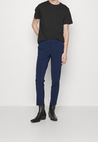 Man som bär svart kortärmad skjorta, marinblå slim fit byxor och svarta ankelstövlar, stående mot en plain vit bakgrund.