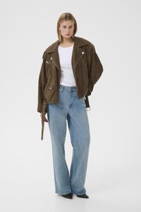 Bruine suède motorjack met zilveren ritsen, brede blauwe spijkerbroek en zwarte schoenen met puntige neus op een effen achtergrond.