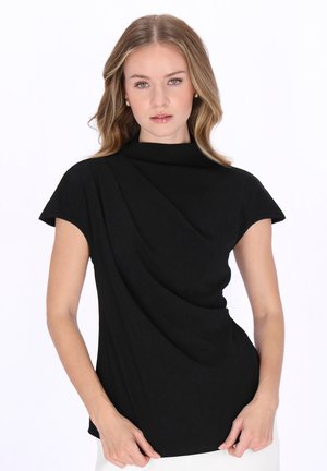 Nuori nainen aaltoilevalla vaalealla hiuksilla, yllään musta draperoitu lyhythihainen paita ja valkoiset housut, seisoo valkoista taustaa vasten.