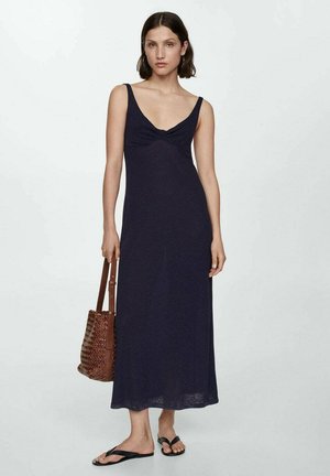 Marineblauwe maxi-jurk met een getailleerde pasvorm, V-halslijn en een twistdetail bij de buste. Gecombineerd met een geweven bruine handtas en zwarte slippers.