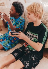 Drenge iført pyjamasæt; den ene i blå med Minecraft-mønstre, den anden i sort med creeper-designs, begge holder spilcontrollere.