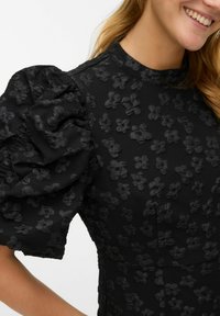 Črna obleka s teksturiranimi cvetličnimi vzorci, napihnjenimi rokavi in visokim ovratnikom. Tkanina je videti lahka s gladkim zaključkom.