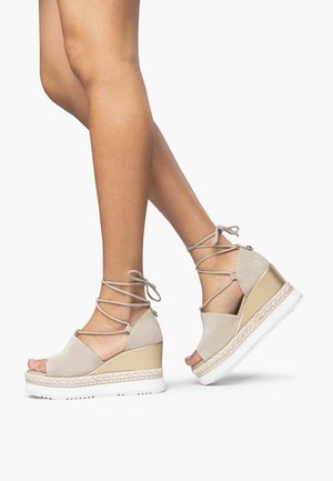 Beige wedge-sandaalit, joissa on mokkanahkainen päällinen, avoin varvas ja nauhoilla suljettava malli. Ominaisuuksiin kuuluu kudottu juuttivälikerros ja valkoinen kumipohja.
