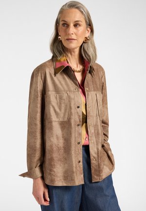 Vrouw met grijs haar die gouden oorringen draagt, een dikke halsketting, een bruine getextureerde blouse met knopen, een kleurrijke blouse en een blauwe broek, staand.
