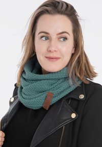 Szydełkowy szal w kolorze turkusowym w kształcie infinity z teksturowanym wzorem i brązową skórzaną metką, noszony z czarną skórzaną kurtką na czarnym swetrze.