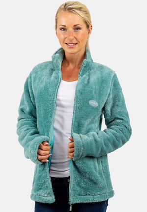 Frau trägt eine türkisfarbene, flauschige Zip-up-Jacke über einem weißen Shirt, hält die Jackenränder mit beiden Händen und lächelt leicht.