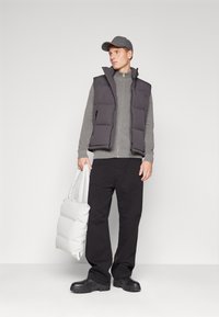 Chaleco de plumón en gris oscuro, combinado con un suéter de cremallera en gris claro y pantalones negros. Accesorio: bolsa de hombro blanca. El modelo lleva una gorra gris.