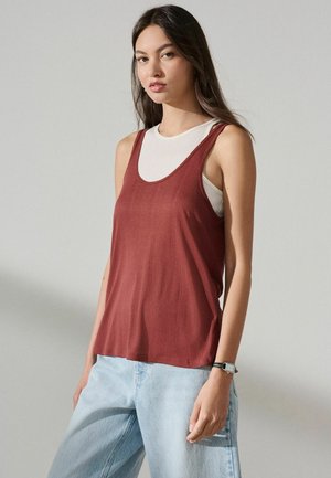 Ung kvinde iført en ærmeløs, rustfarvet top over en hvid tanktop, lyseblå jeans og et sort armbåndsur, stående foran en ensfarvet baggrund.