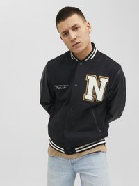 Zwarte varsityjas met gouden en witte letter "N" op de borst, gestreepte manchetten en zijzakken, gecombineerd met een beige shirt en lichte jeans.