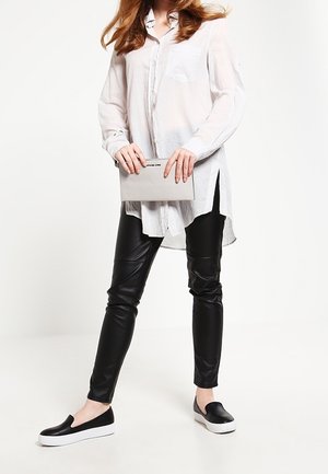 Žena v bílé dlouhé košili, černých kalhotách z umělé kůže a černých nazouvacích botách držící světle šedé psaníčko.