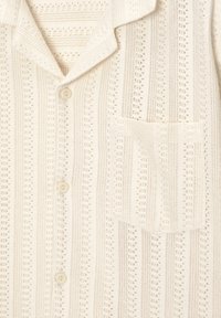 Chemise tricotée couleur crème avec motif ajouré vertical, boutons à l'avant, col et poche poitrine sur le côté droit.