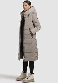 Pikk beež puffjakker, millel on chevron-muster, kõrge krae, ees zipp-taskud ja sildike, mis ripub tõmbluku küljes. Kanti mustade retuuside peal.