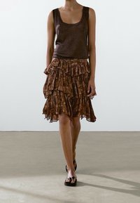 Top marrone senza maniche abbinato a una gonna a strati con volant caratterizzata da un motivo tie-dye. La modella indossa ciabatte nere. Sfondo con pavimento liscio.