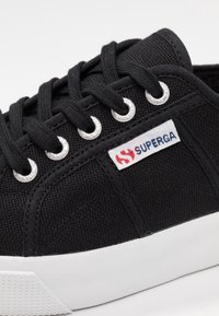 Svart kanvas sneakers med svarta snören, silverfärgade öglor, vit gummisula och en vit etikett med den röda och blå Superga-logotypen.