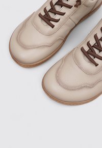 Beige leather sneakers with a textured finish, featuring brown laces, round toe, and contrasting rubber sole.