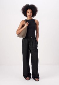 Top de alças preto com textura canelada, calças largas pretas com cordão e sandálias pretas. Segurando uma mala curva de tom castanho claro. Design simples.