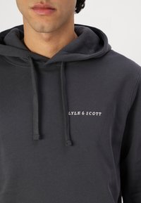 Temno siva kapuca z kenguru žepom, vezicami in belim tekstom "LYLE & SCOTT" na levi strani. Bombažni material s mehko teksturo.