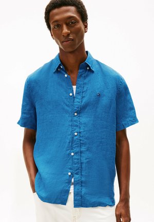 Hombre con camisa de manga corta de botones de color azul brillante sobre una camiseta blanca, de pie con las manos en los bolsillos contra un fondo blanco liso.