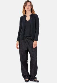Schwarzer Cardigan mit Rundhalsausschnitt und Knopfverschluss, darüber ein schwarzes V-Ausschnitt-Oberteil. Locker sitzende schwarze Hose mit elastischem Bund und Taschen.
