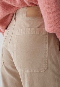 Pantalon en velours côtelé beige à la texture lisse, doté de deux poches arrière et d'un patch en cuir sur la ceinture embossé du mot "OPUS".