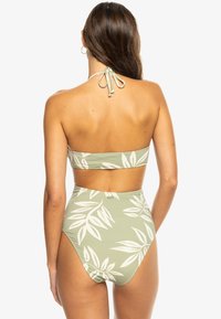 Hoog uitgesneden bikini in lichtgroen met grote witte bladrandpatronen. Halternekontwerp met een strik aan de achterkant en een textuurstof.