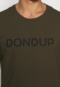 Camiseta verde oliva de tejido suave con letras negras que dicen "DON DUP", con cuello redondo y mangas cortas.