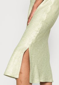 Zelené šaty s vzorem a přiléhavým střihem, které mají jemnou mřížkovou texturu a boční rozparek pro snadný pohyb.