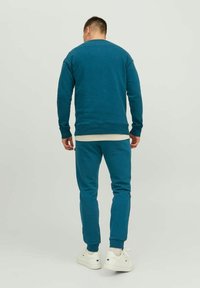 Teal sweatshirt en bijpassende joggingsbroek van zacht materiaal. Voorzien van ribbels bij de manchetten en de tailleband. Draagt met witte sneakers.