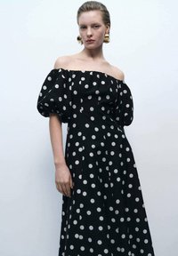 Rochie neagră cu umeri descoperiți, cu buline mari albe, mâneci bufante și o siluetă fluidă, confecționată dintr-un material ușor.