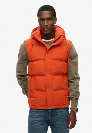 Padded orange vest with a hood, featuring snap buttons and drawstrings. Worn over a beige sweater, paired with blue jeans.