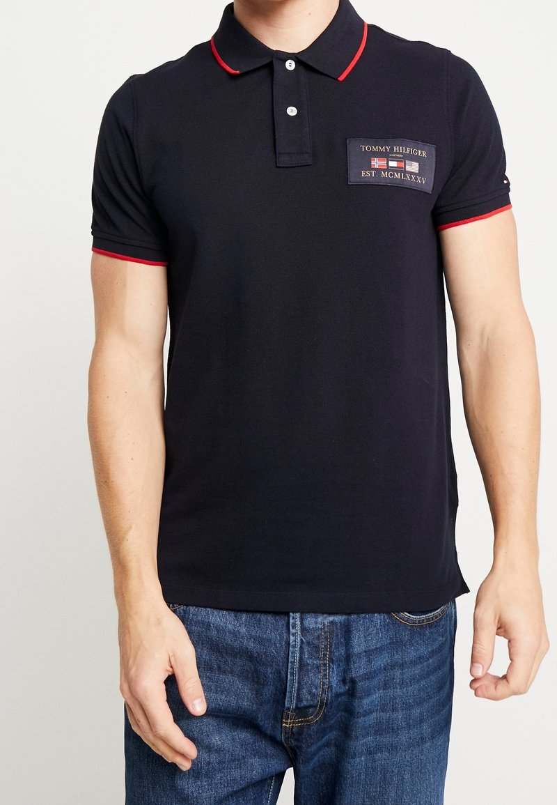 Homme portant un polo bleu marine avec une bordure rouge et un écusson du logo Tommy Hilfiger sur la poitrine, associé à un jean bleu.