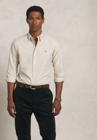 Beige collared shirt with a small logo, paired with dark green trousers and a brown belt. The shirt has rolled-up sleeves and a smooth texture.