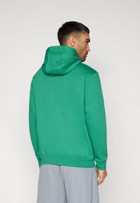 Πράσινο φούτερ με κουκούλα και κορδόνι, υφή υφάσματος, ριμπ μανσέτες και τελείωμα. Αγκαζέ από πίσω, συνδυασμένο με γκρι σορτς.