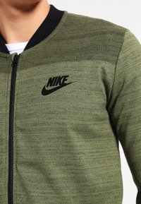 Olive green Nike bomber jacket with textured fabric, black collar, and zipper. Features prominent black logo on left chest.