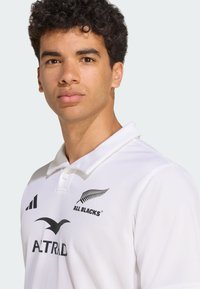 Bílé ragbyové tričko s klasickým límečkem, texturovanou tkaninou a černými logy "All Blacks," "Altrad" a třemi pruhy na rameni.